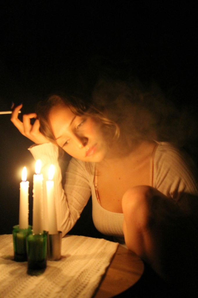 A woman smokes pensively by candlelit table, surrounded by darkness.